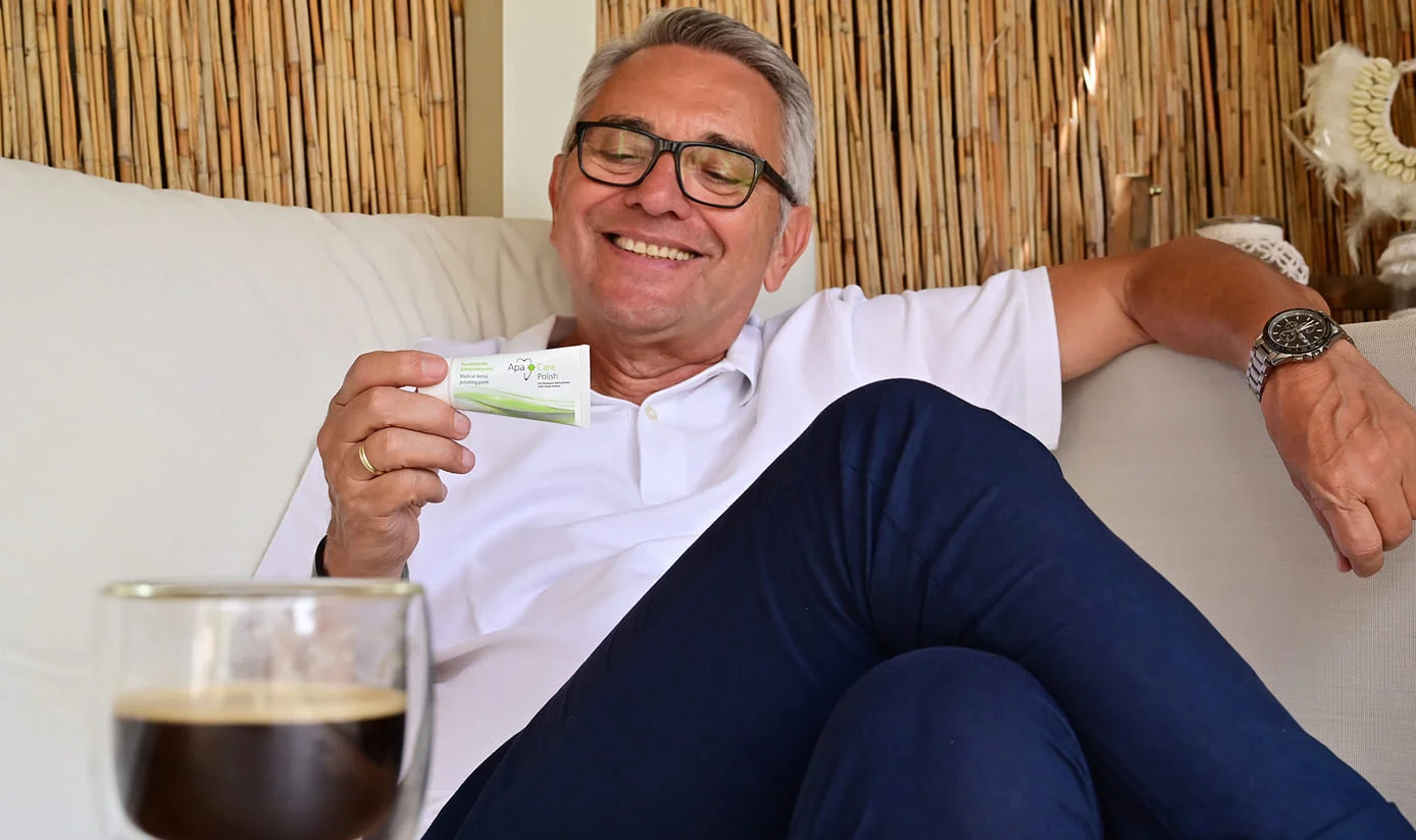 Mann hält ApaCare Zahn-Polierpaste in der Hand, vor ihm steht ein Glas mit schwarzem Kaffee